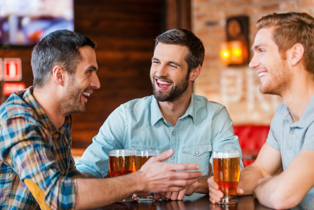 Three happy young men in casual wear talking and drinking beer while sitting in bar together