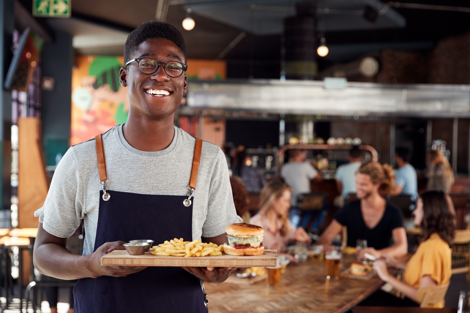 Portrait Of Waiter Serving Food To Customers In Busy Bar Restaurant
